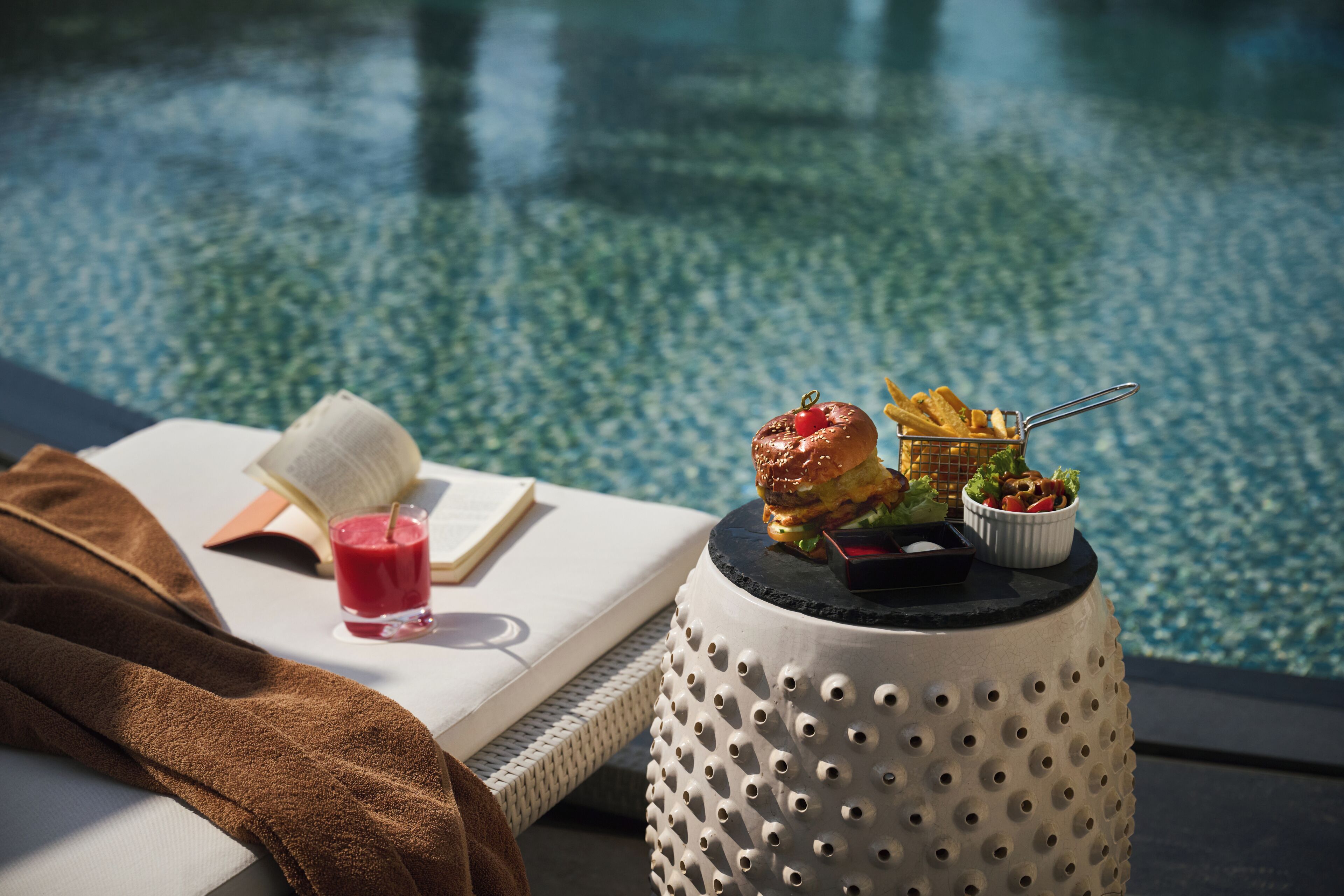 Escena junto a la piscina con un libro, una bebida, una hamburguesa, papas fritas y una ensalada sobre una mesa.