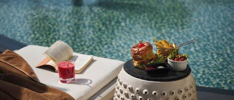 Escena junto a la piscina con un libro, una bebida, una hamburguesa, papas fritas y una ensalada sobre una mesa.