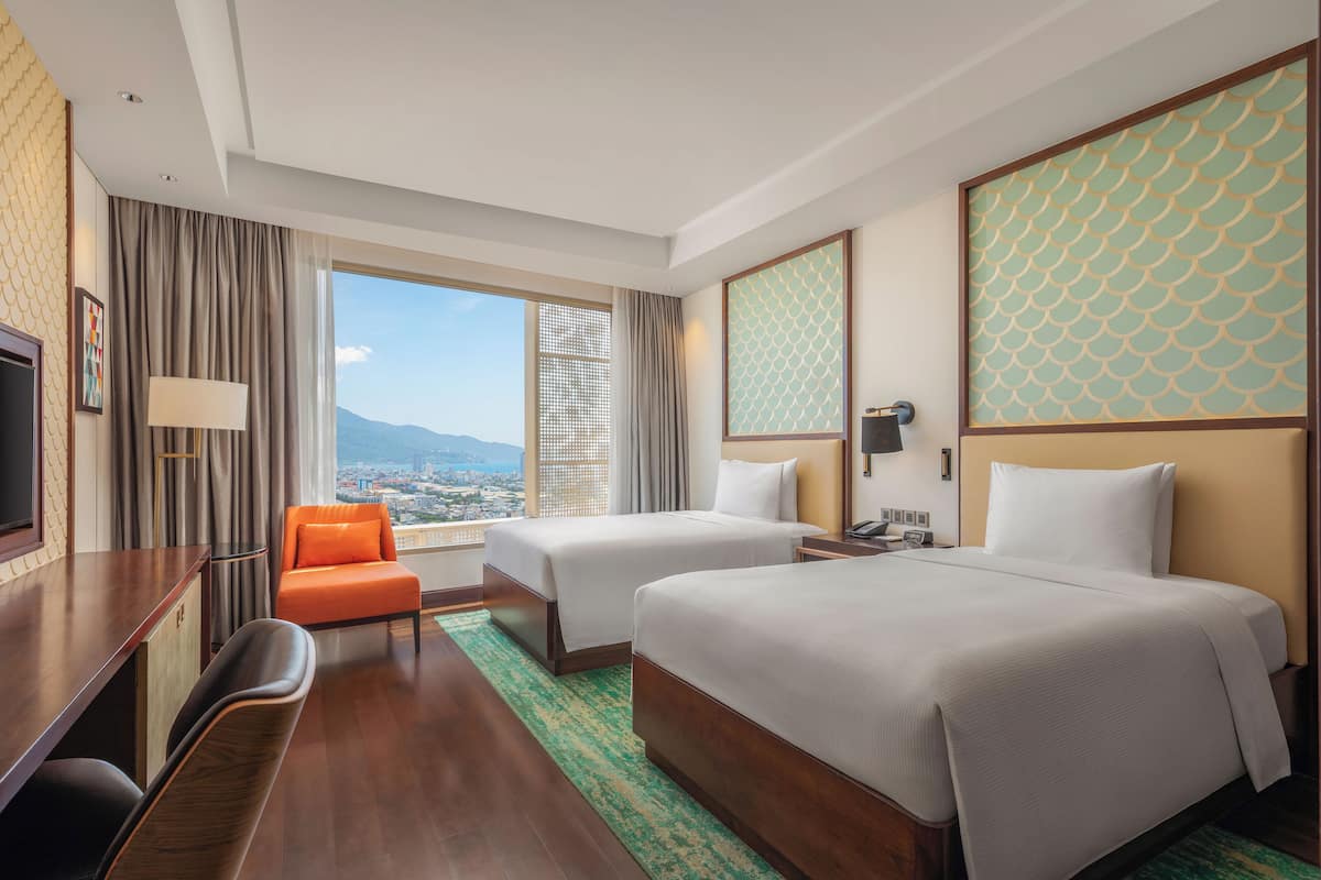 A hotel room with two beds, a desk, a chair, a TV, and a view of the city.