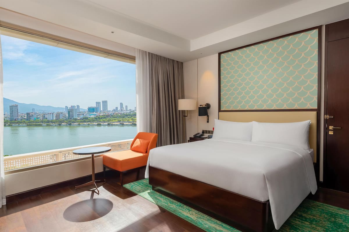 A hotel room with a bed, a chair, a table, a lamp, and a view of a cityscape and water.