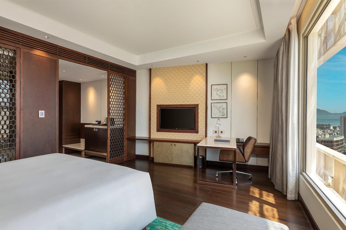 A hotel room with a bed, desk, chair, TV, and a view of the city.