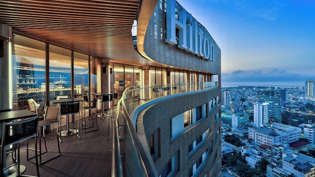 Un hotel moderno de gran altura, con un bar en la azotea que cuenta con mesas y sillas, amplias ventanas y vistas panorámicas de la ciudad.