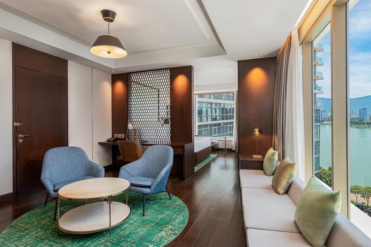 A modern hotel room with a sofa, two armchairs, a round table, a patterned rug, a decorative wall, and a view of a cityscape through large windows.