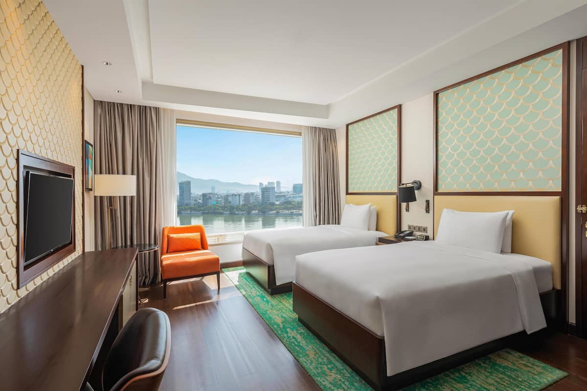 A hotel room with two beds, a desk with a TV, a chair, a view of a cityscape, and a large window with curtains.