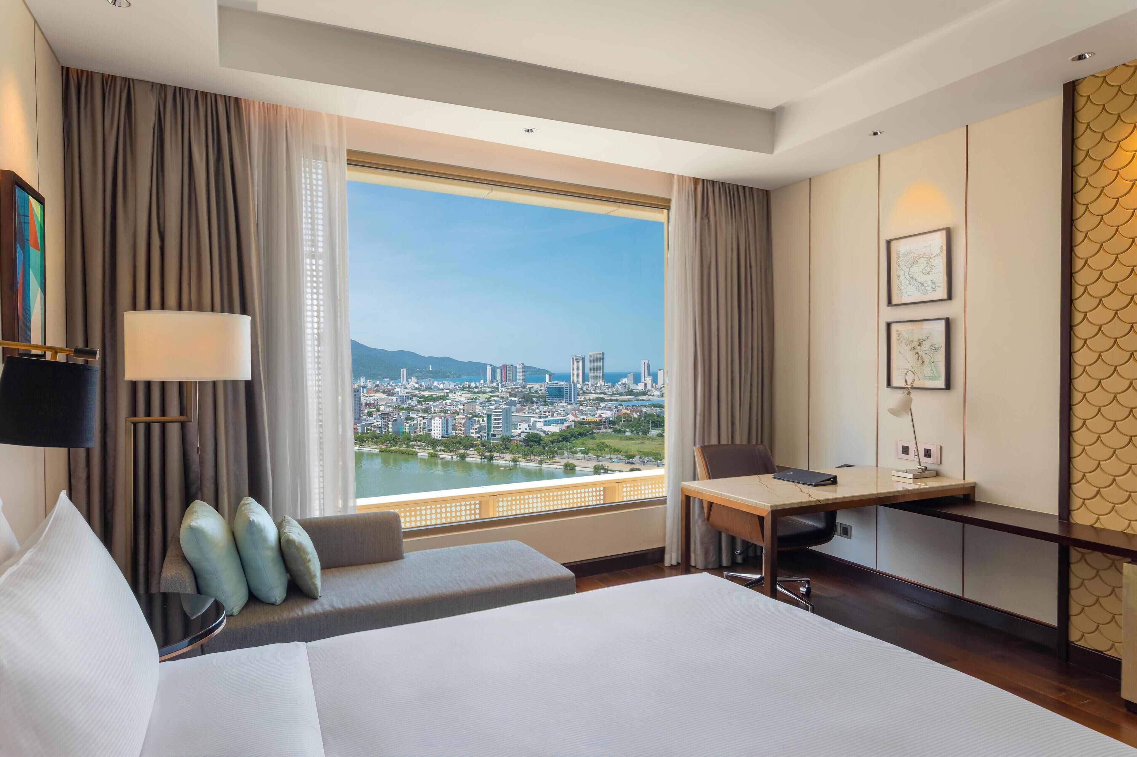 A hotel room with a large bed, a desk, a view of a cityscape, and a sofa.