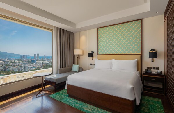 A hotel room with a large bed, a small table, a sofa, a nightstand, and a view of the city. - Hilton Da Nang (Da Nang)