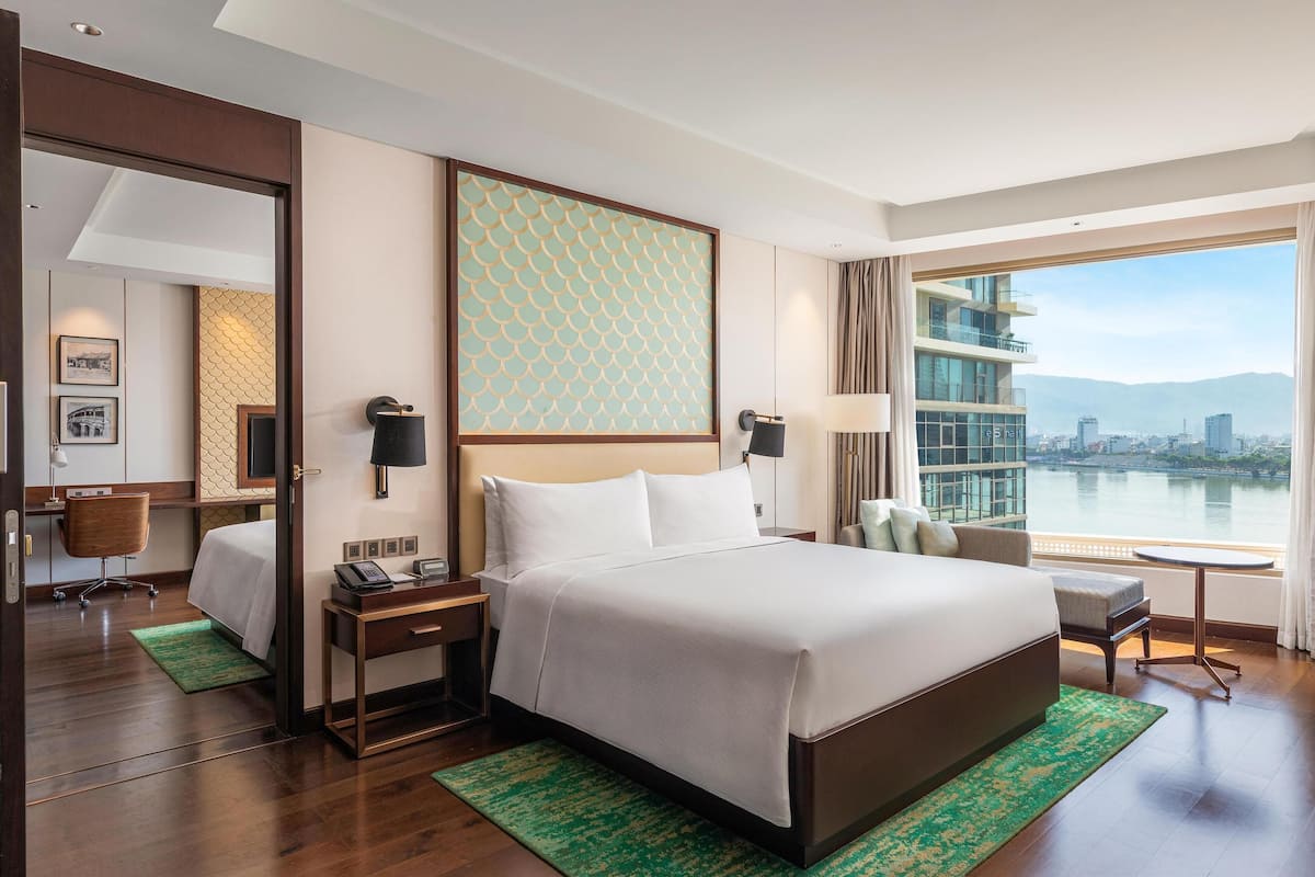 A hotel room with a large bed, a desk, a chair, a nightstand, a lamp, and a view of a cityscape through a large window.