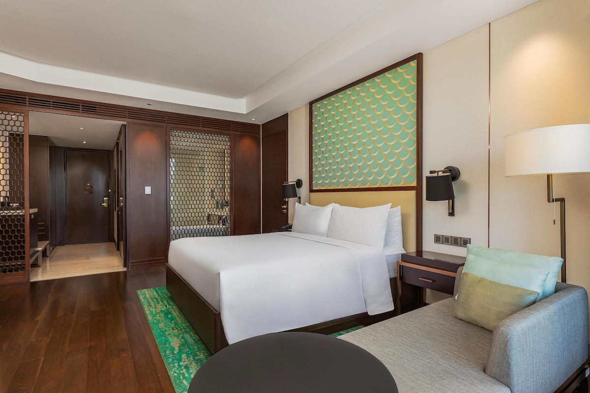 A hotel room with a large bed, a desk with a lamp, a chair, and a decorative wall panel.