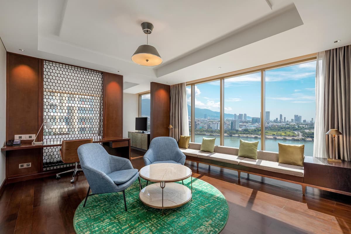 A modern hotel room with a large window offering a city view, a cozy seating area with a sofa and chairs, a small round table, and a patterned wall.