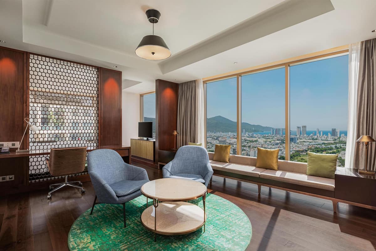 A modern hotel room with a large window, a round table, two armchairs, a sofa, and a decorative wall.