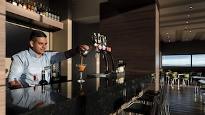 A bar area with a bartender pouring a drink, a selection of bottles on a shelf, and a view of the outside through large windows. - Hampton by Hilton Bogota Airport (Bogotá)