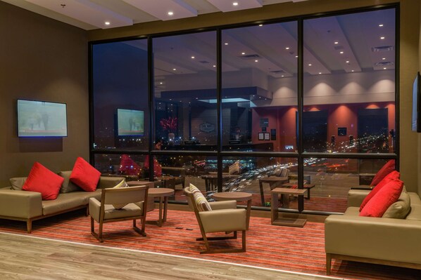 A modern hotel lobby with a large window offering a city view at night. There are sofas, armchairs, and small tables arranged for guests.