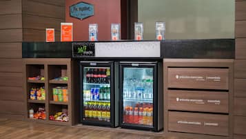 Un mostrador de bar de aperitivos con dos refrigeradores llenos de bebidas y aperitivos, un cartel que indica disponibilidad de aperitivos las 24 horas, y un cartel sobre los refrigeradores que dice "Hamptons by Hilton".