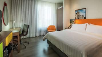 A hotel room with a bed, a desk with a chair, a large window with curtains, and a painting on the wall.