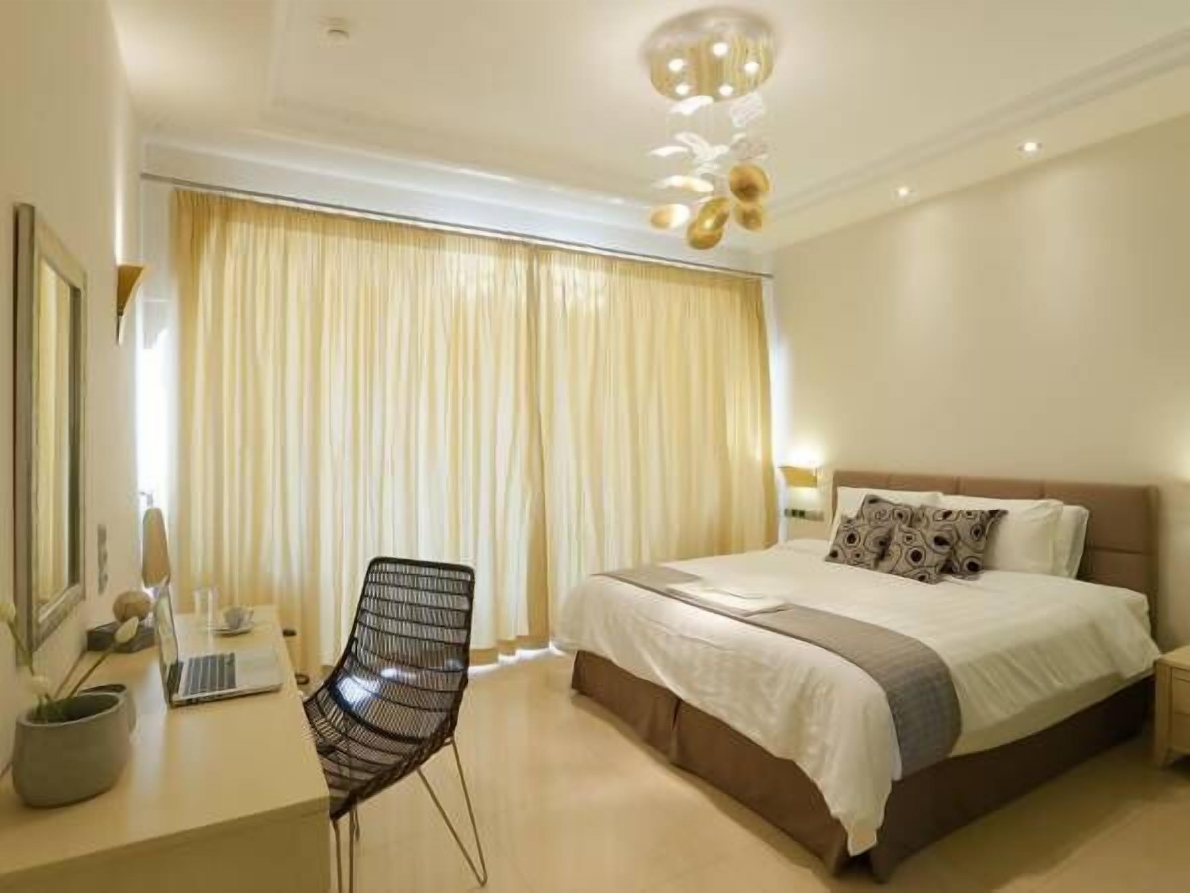 Habitación de hotel con una cama grande, un escritorio con una computadora portátil, una silla y un ventanal con cortinas.