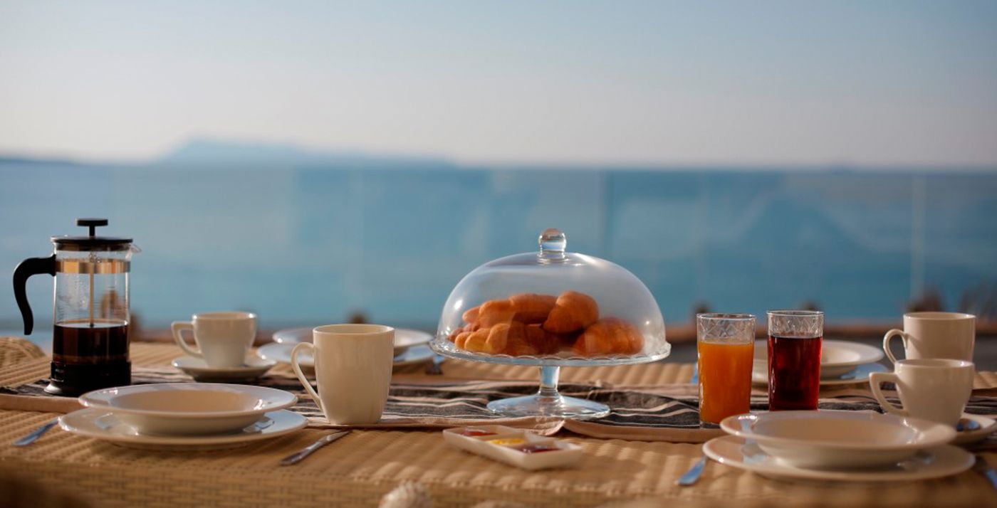 Ein Frühstückstisch mit Kaffeekanne, Orangensaft im Glas und einer Auswahl an Gebäck, mit Blick auf das Meer.