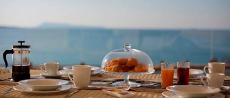 Ein Frühstückstisch mit Kaffeekanne, Orangensaft im Glas und einer Auswahl an Gebäck, mit Blick auf das Meer.