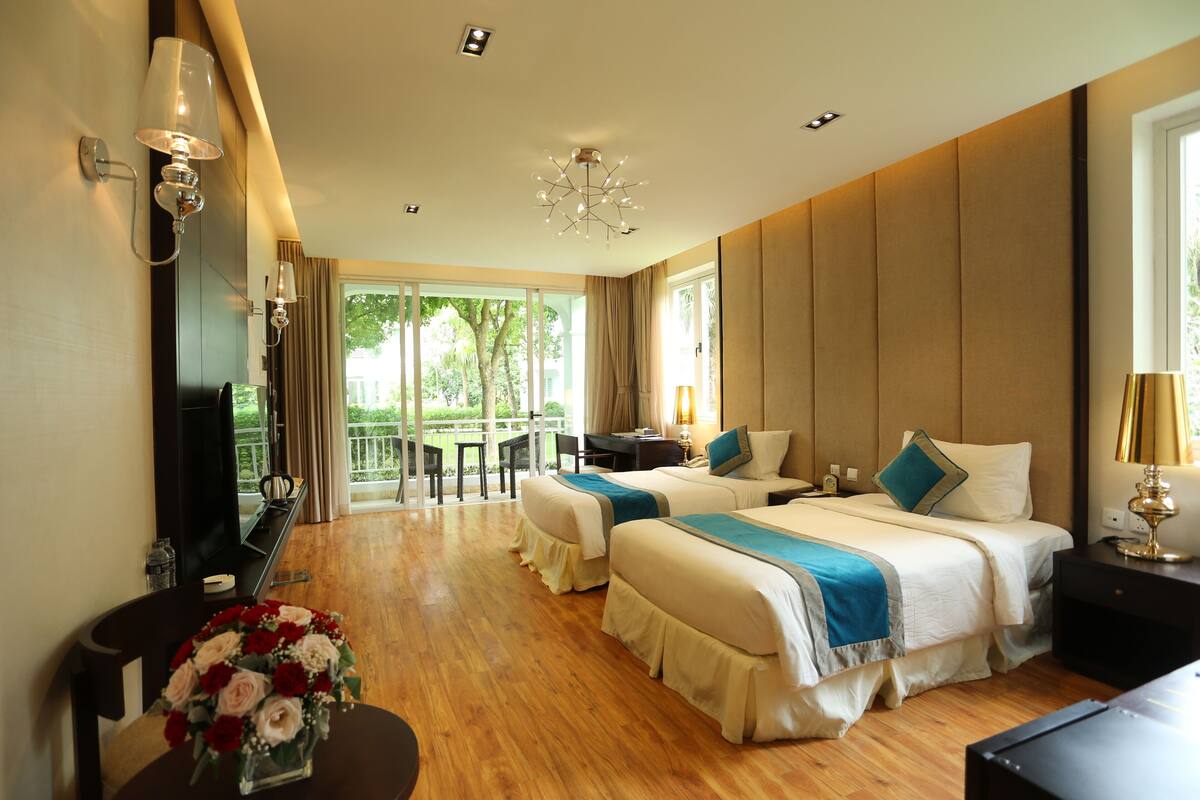 A hotel room with two beds, a desk, a chair, a TV, and a balcony with a view of trees.