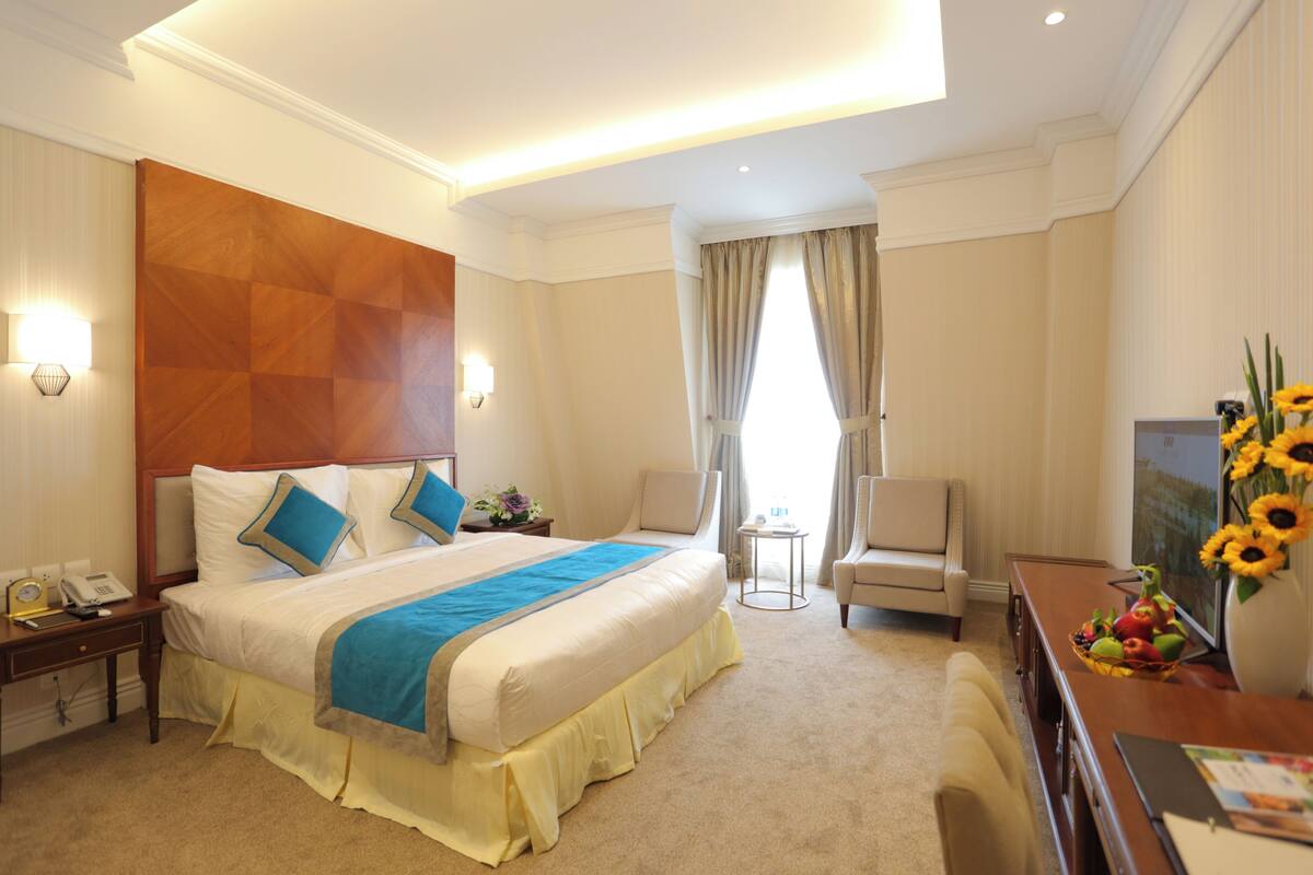 A hotel room with a large bed, a desk with a vase of sunflowers, a chair, and a small table with fruit.