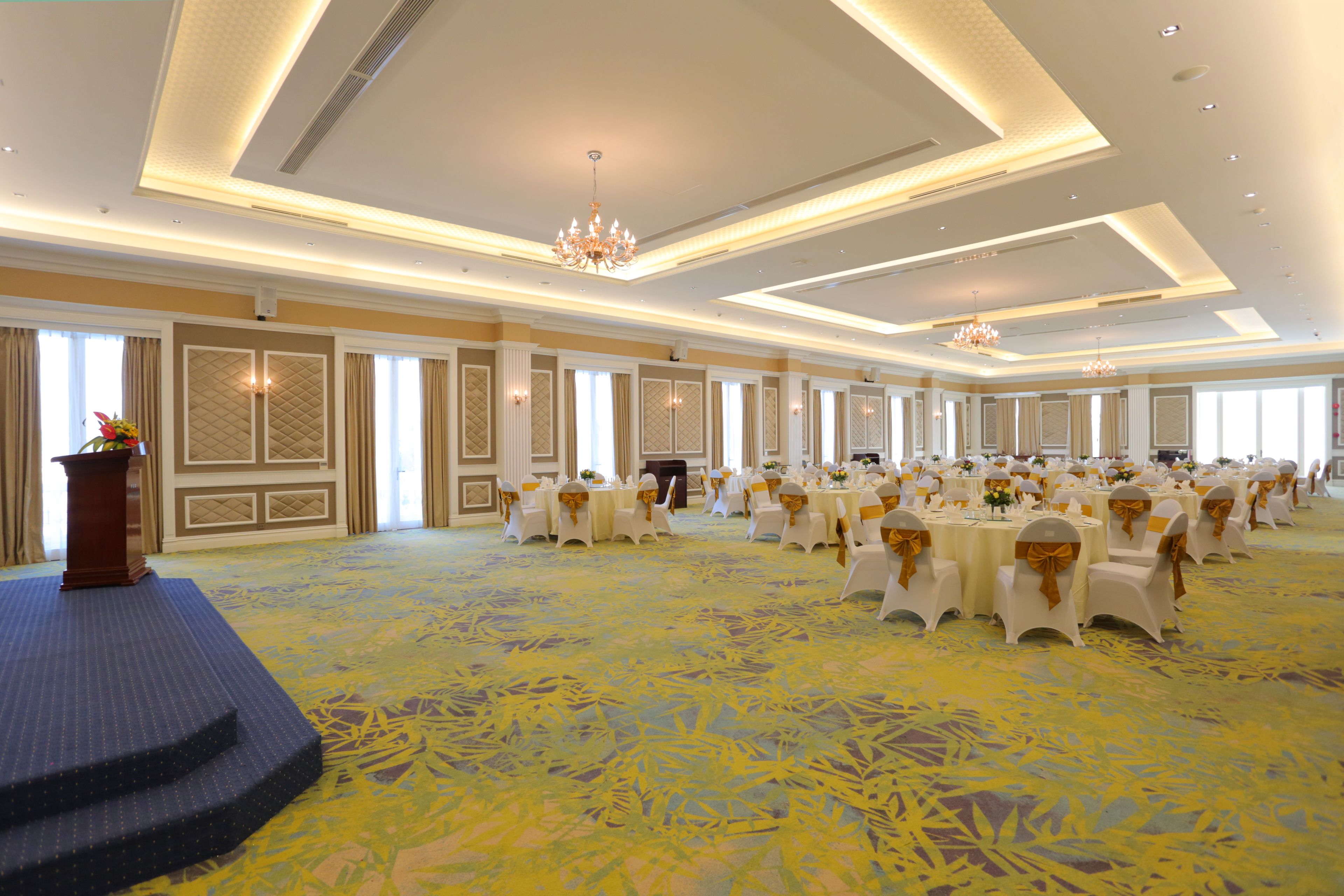A large banquet hall with rows of round tables, a stage with a podium, and chandeliers.