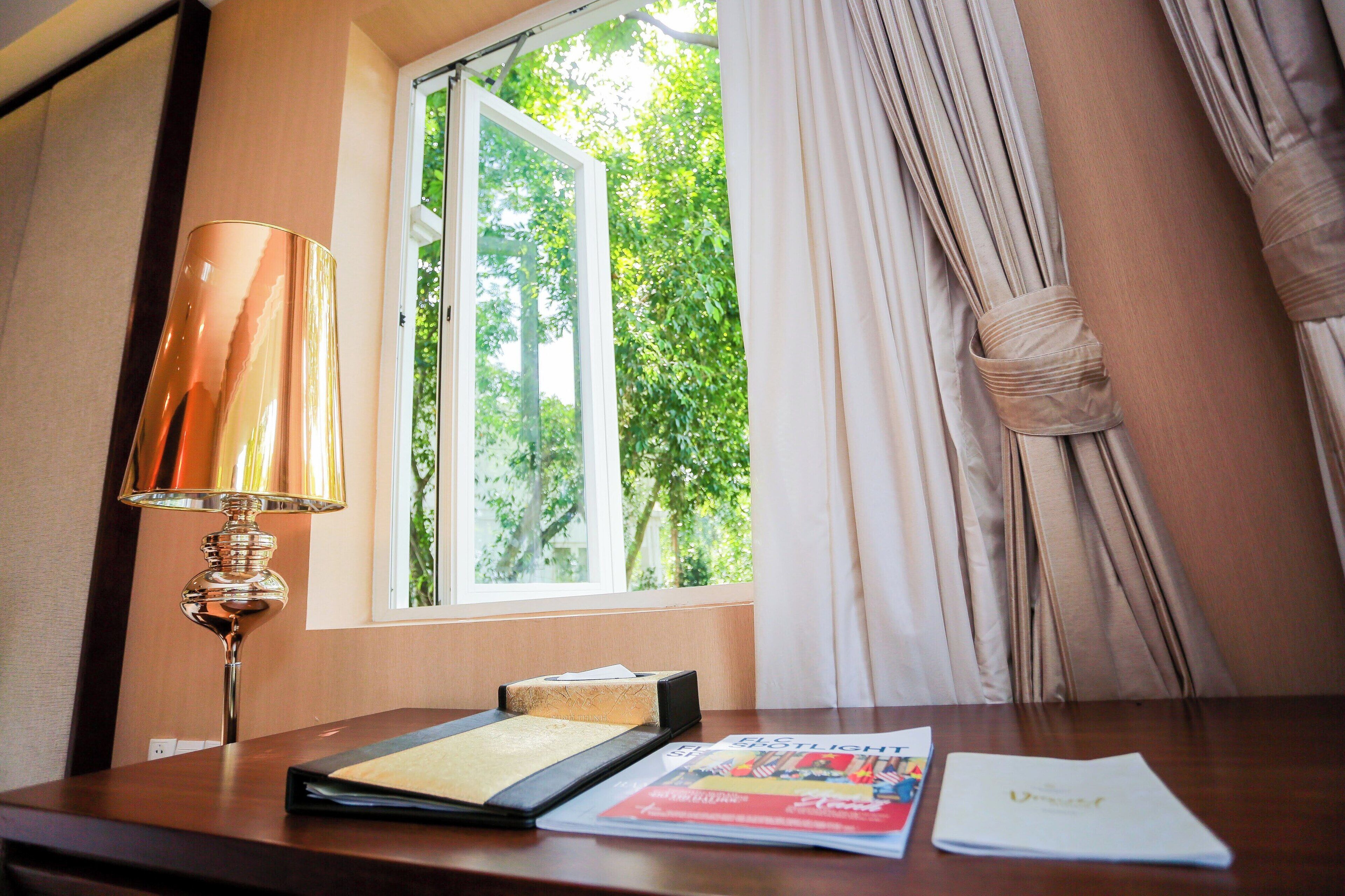 Quarto de hotel com uma secretária de madeira, uma luminária, uma janela com cortinas e vista para áreas verdes.