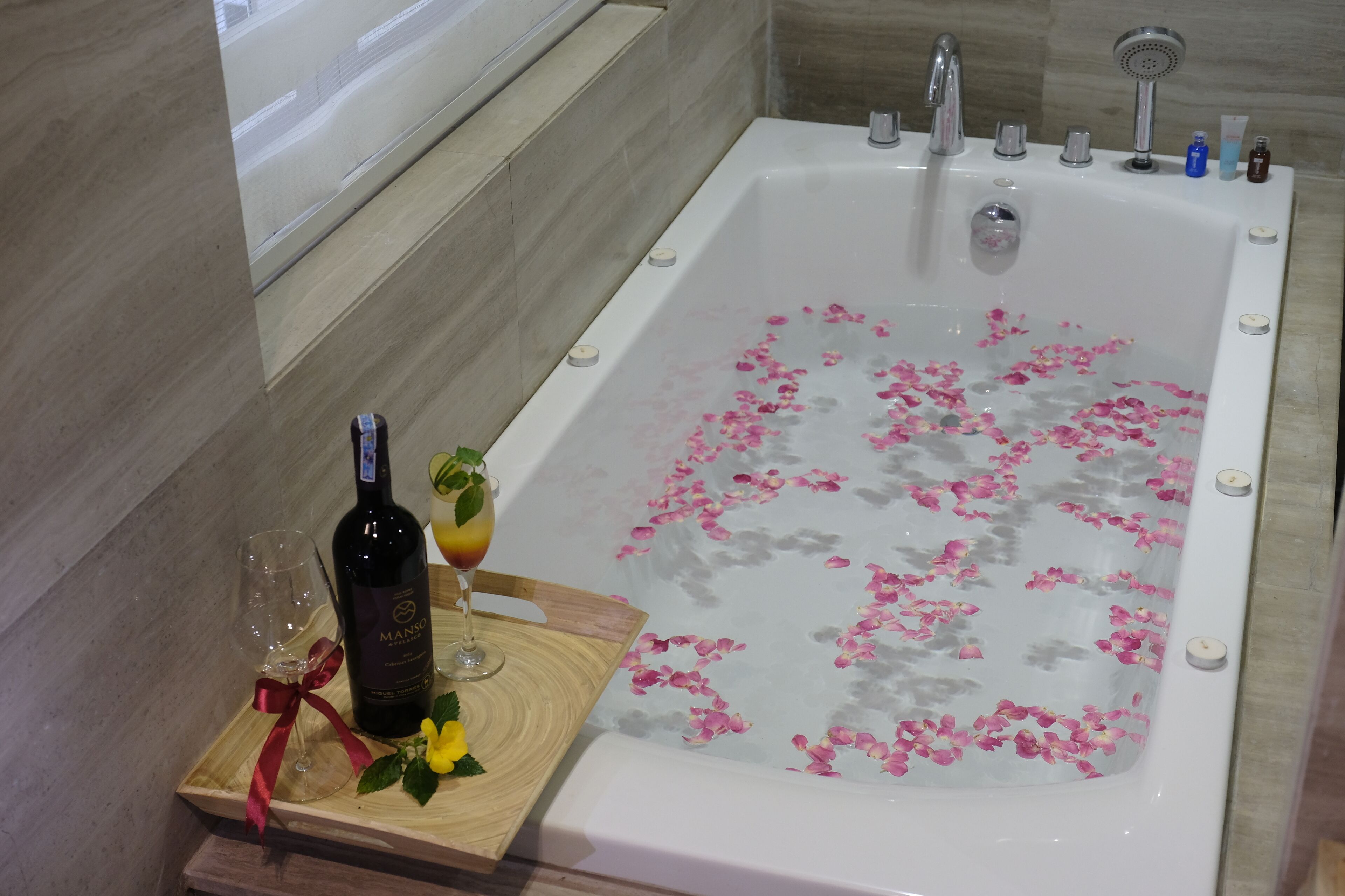 Eine Badewanne gefüllt mit Wasser und Rosenblüten, eine Weinflasche und ein Glas auf einem Holztablett.