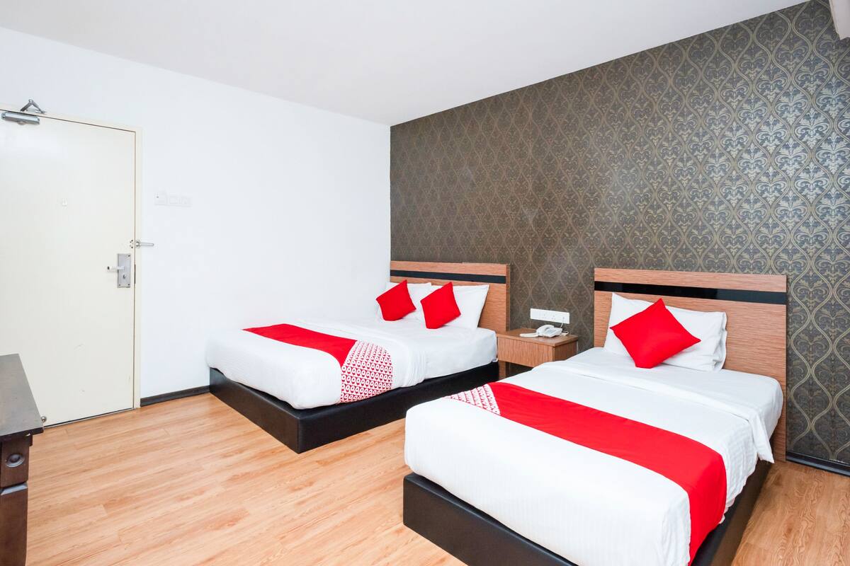 A hotel room with two beds, each with white bedding and red pillows, a wooden floor, and a patterned wallpaper on the wall.
