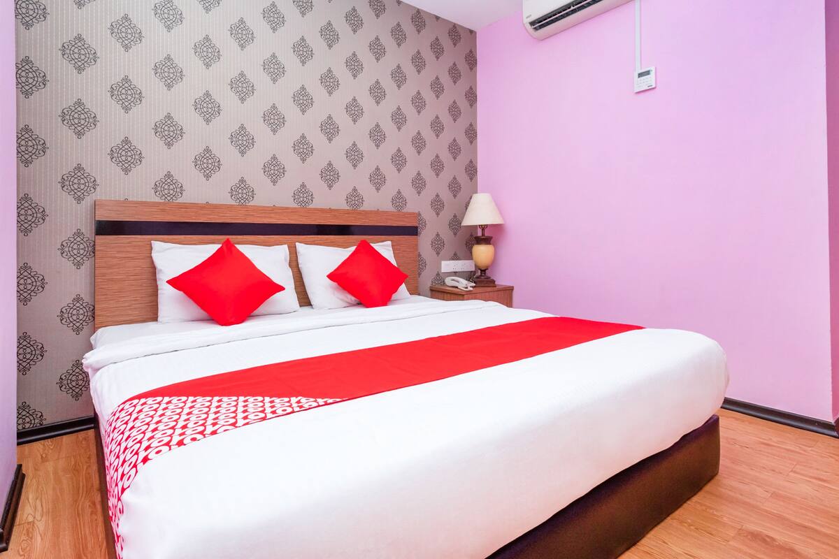A hotel room with a large bed, a wooden headboard, a bedside table with a lamp, and an air conditioning unit on the wall.