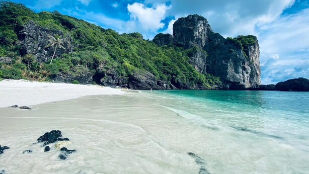 Une plage à l’eau turquoise et limpide, bordée de sable blanc, avec, en arrière-plan, des collines verdoyantes.