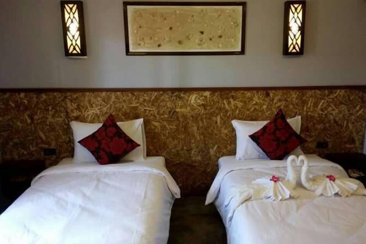 A hotel room with two beds, each with white bedding and red pillows. There are wall-mounted light fixtures above the beds and a framed artwork on the wall.