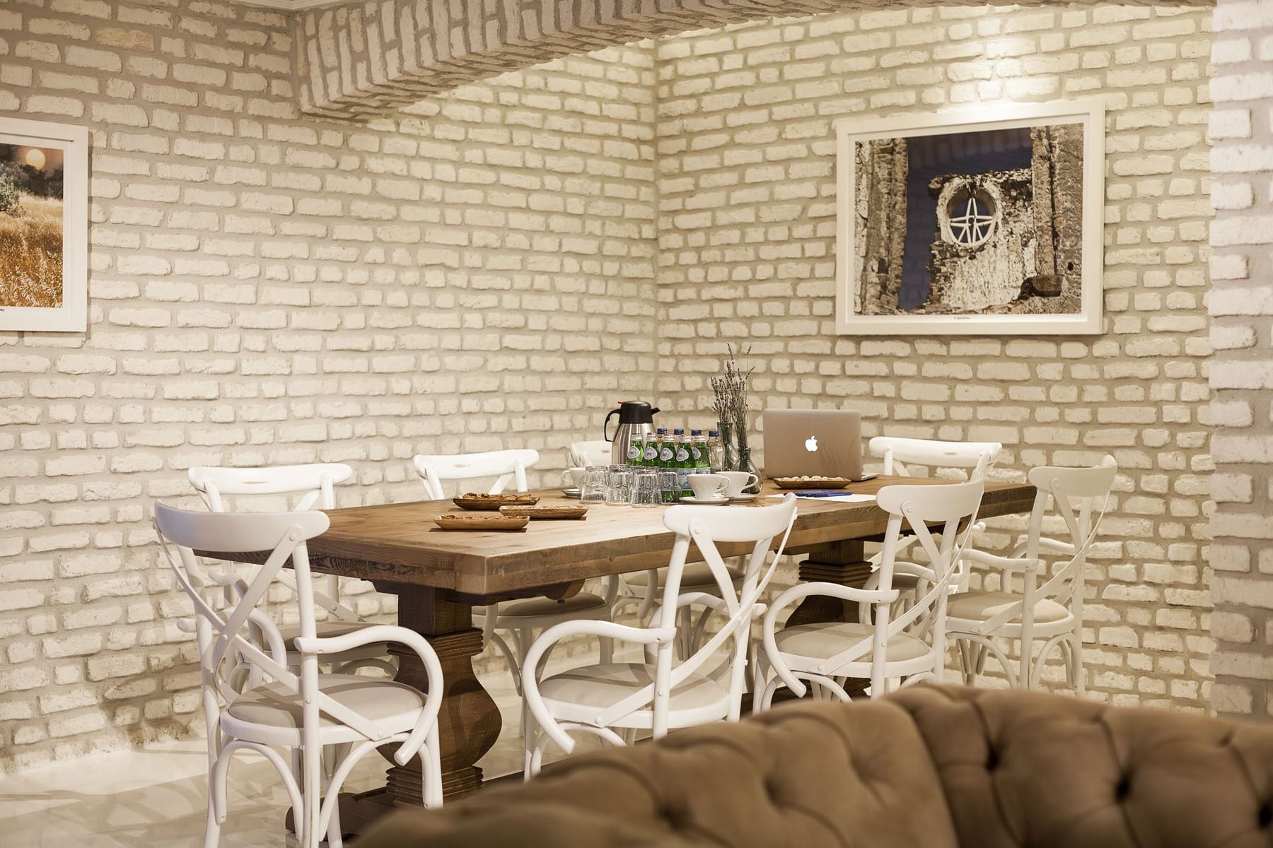 A dining area with a brick wall, a wooden table, white chairs, a laptop, and a coffee maker.