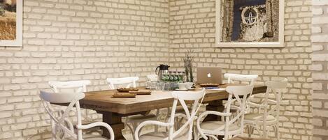 A dining area with a brick wall, a wooden table, white chairs, a laptop, and a coffee maker.