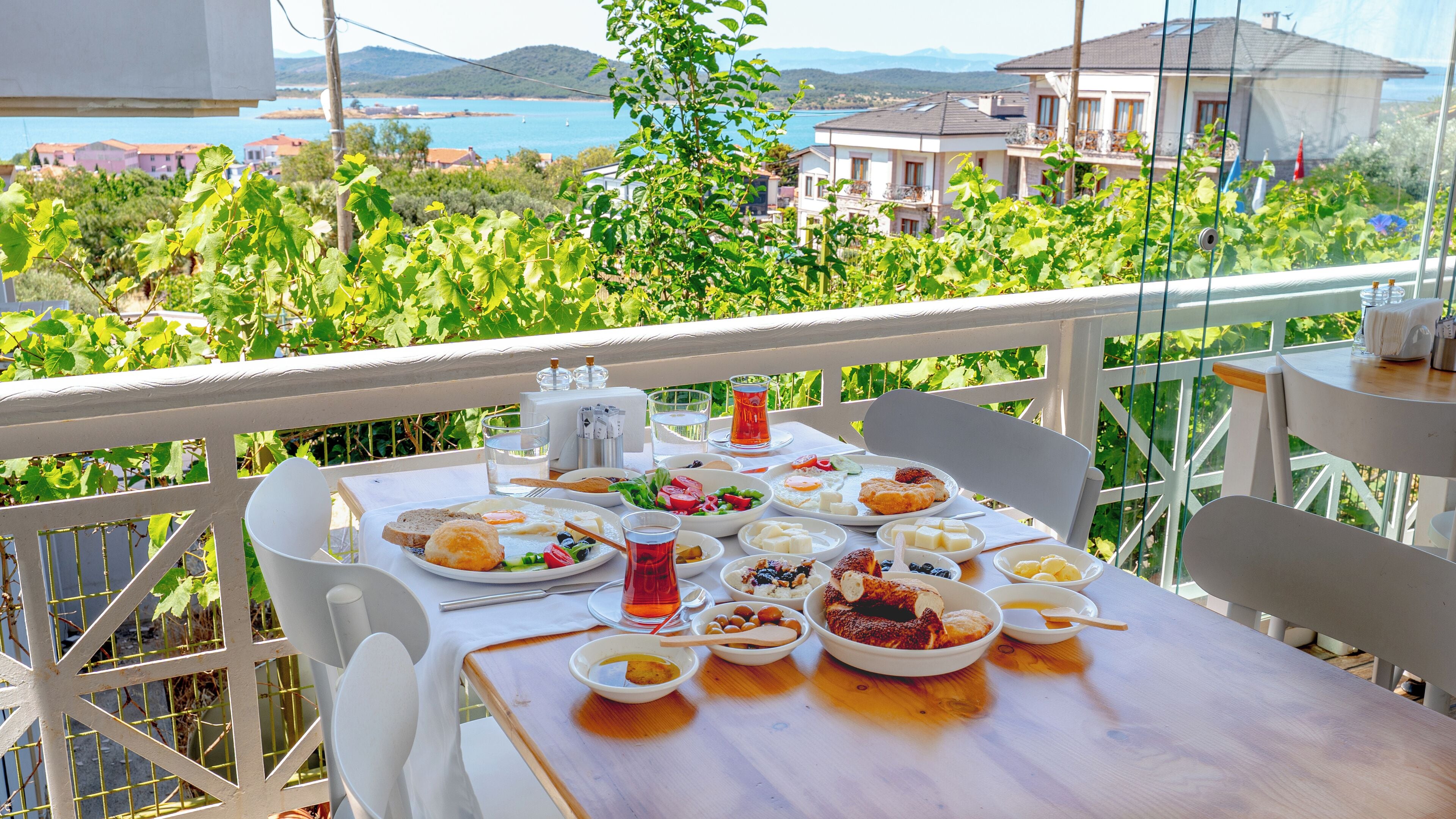 A table set with various dishes and drinks on a balcony overlooking a scenic view of a coastal area with houses and greenery.