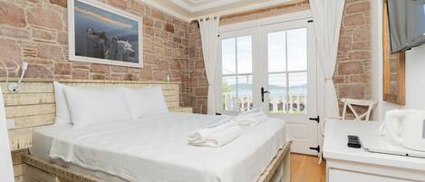 Une chambre avec un mur en pierre, un lit recouvert de draps blancs, un tableau représentant des chevaux, un téléviseur et une vue sur la mer, visible à travers une porte.