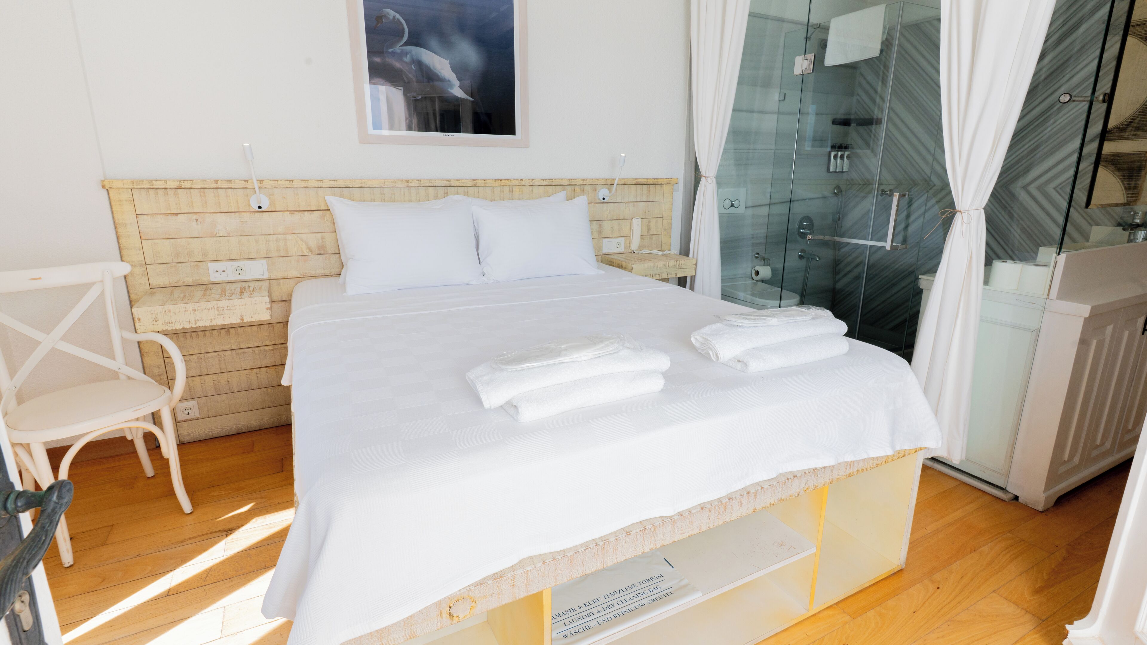 A neatly made bed with white linens and towels, a wooden headboard, a white chair, and a glass shower enclosure.