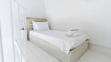 Un lit simple avec du linge de lit blanc et une tête de lit en bois, dans une pièce aux murs et au sol blancs.