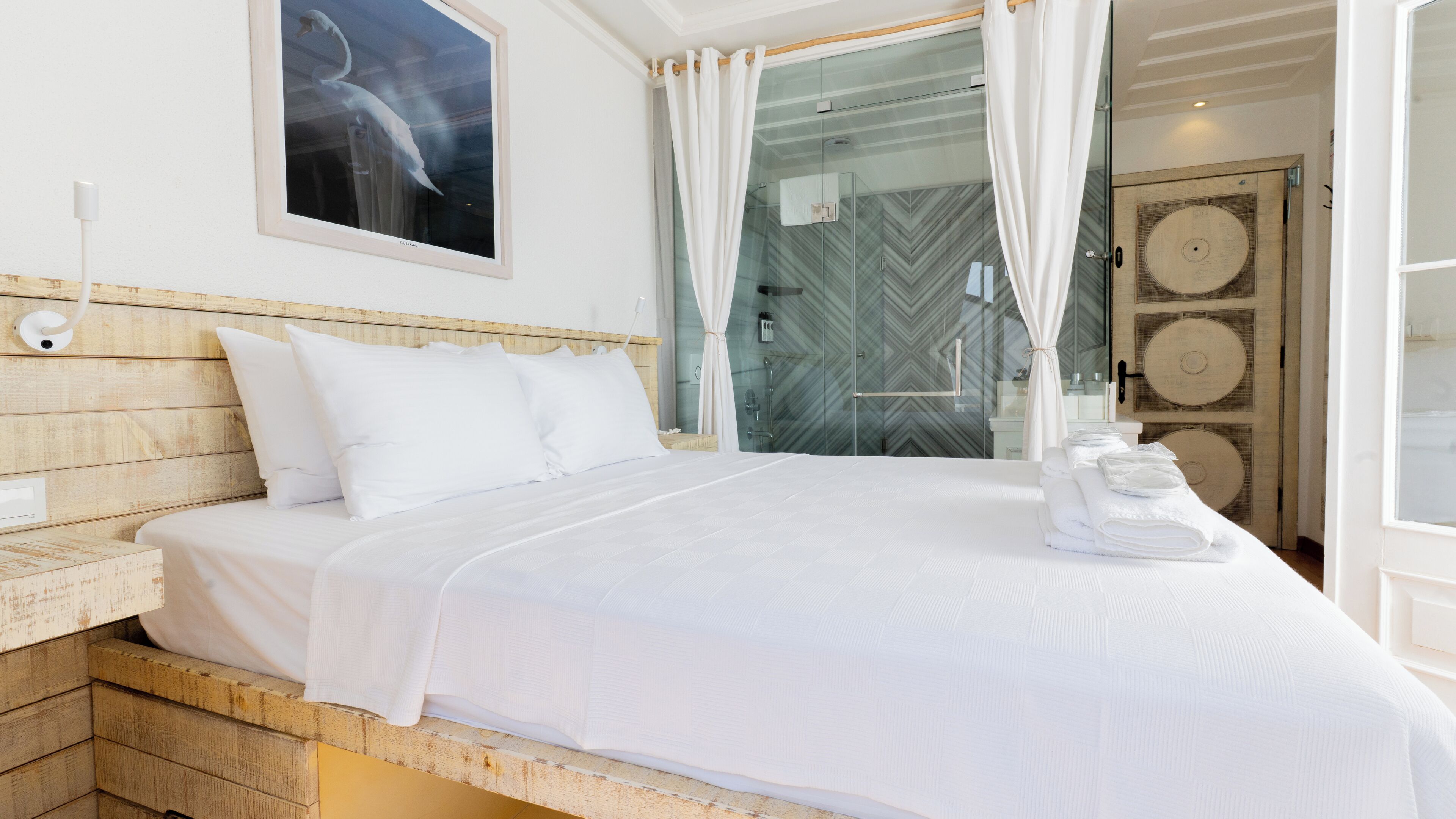 A neatly made bed with white linens, a wooden headboard, and a glass shower enclosure with a herringbone-patterned wall.
