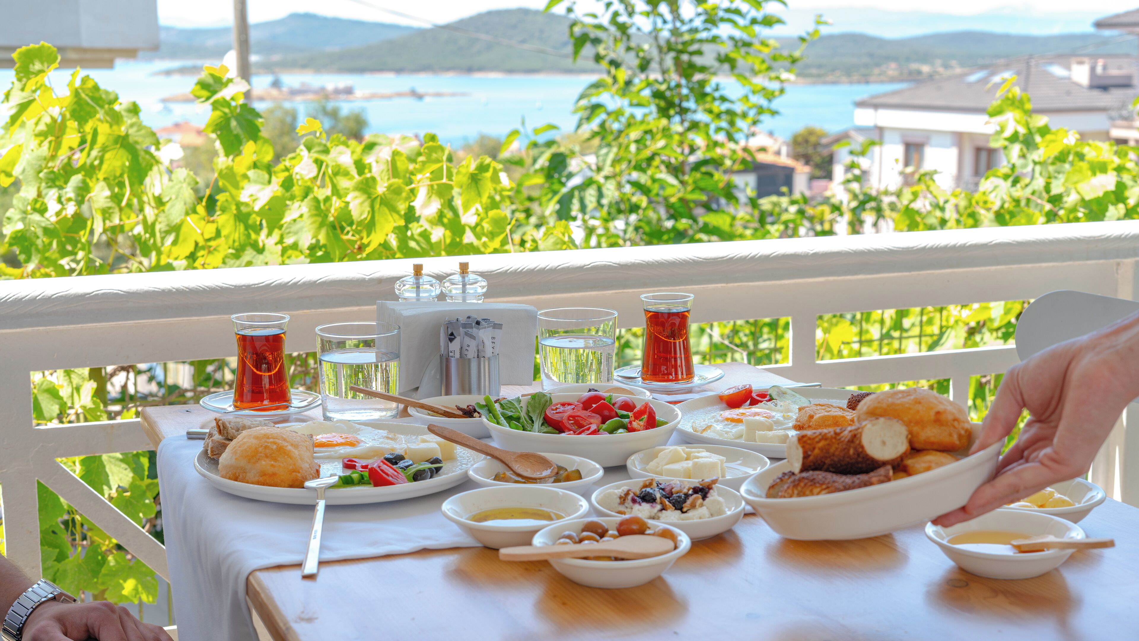 A table set with various dishes and drinks, overlooking a scenic view of a body of water and greenery.