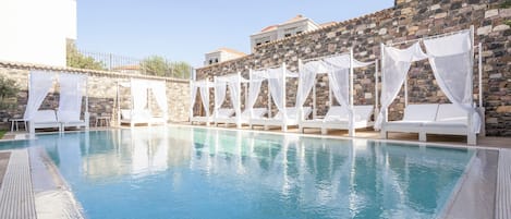 Una piscina con sillas de descanso blancas y cortinas translúcidas, rodeada de muros de piedra y un edificio blanco.