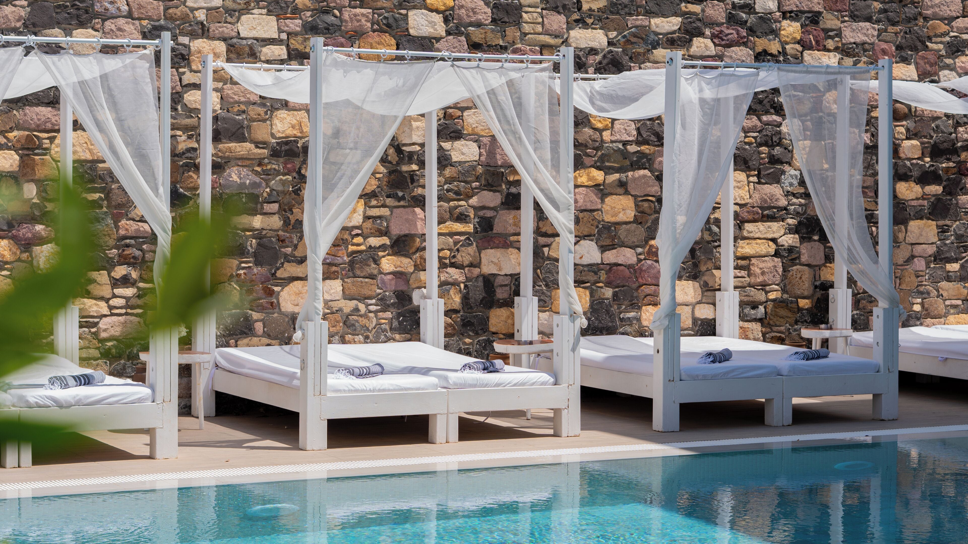 A poolside area with four white beds, each with a canopy, set against a stone wall.