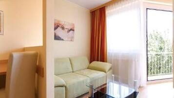 Habitación de hotel con kitchenette, sofá, televisor y balcón con cortinas.