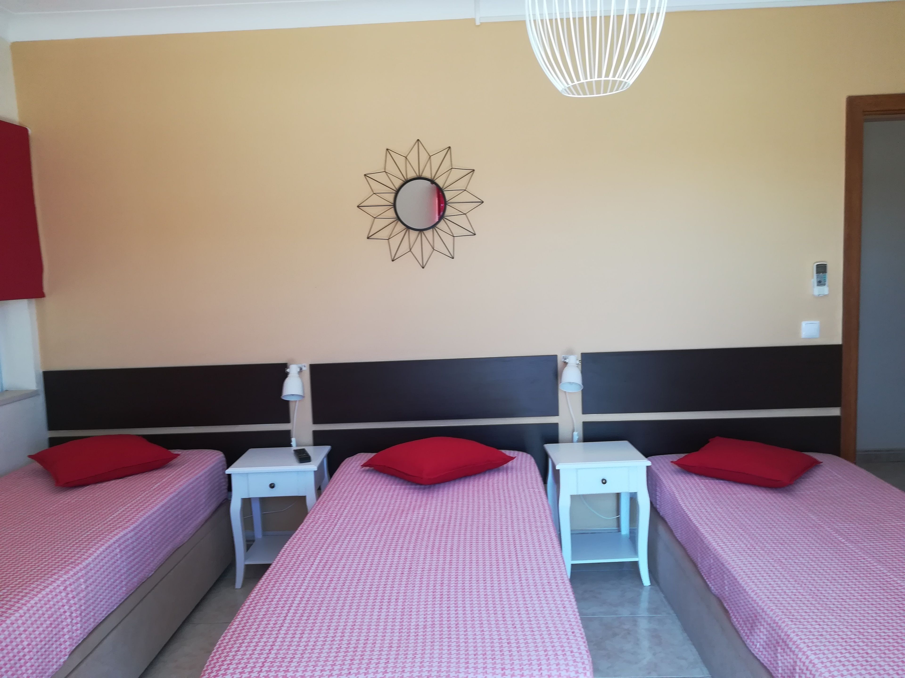 Habitación de hotel con tres camas individuales, cada una con una almohada roja y una mesita de noche blanca. Se aprecia un espejo montado en la pared con diseño geométrico, una lámpara colgante blanca y un enchufe.