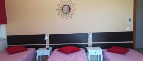 Habitación de hotel con tres camas individuales, cada una con una almohada roja y una mesita de noche blanca. Se aprecia un espejo montado en la pared con diseño geométrico, una lámpara colgante blanca y un enchufe.
