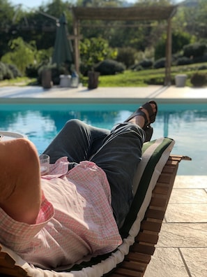 Una persona recostada en una silla de madera junto a una piscina, con una pérgola y follaje al fondo.