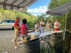Un grupo de personas alrededor de una mesa con equipo de cocina, al aire libre con un coche y follaje en el fondo.