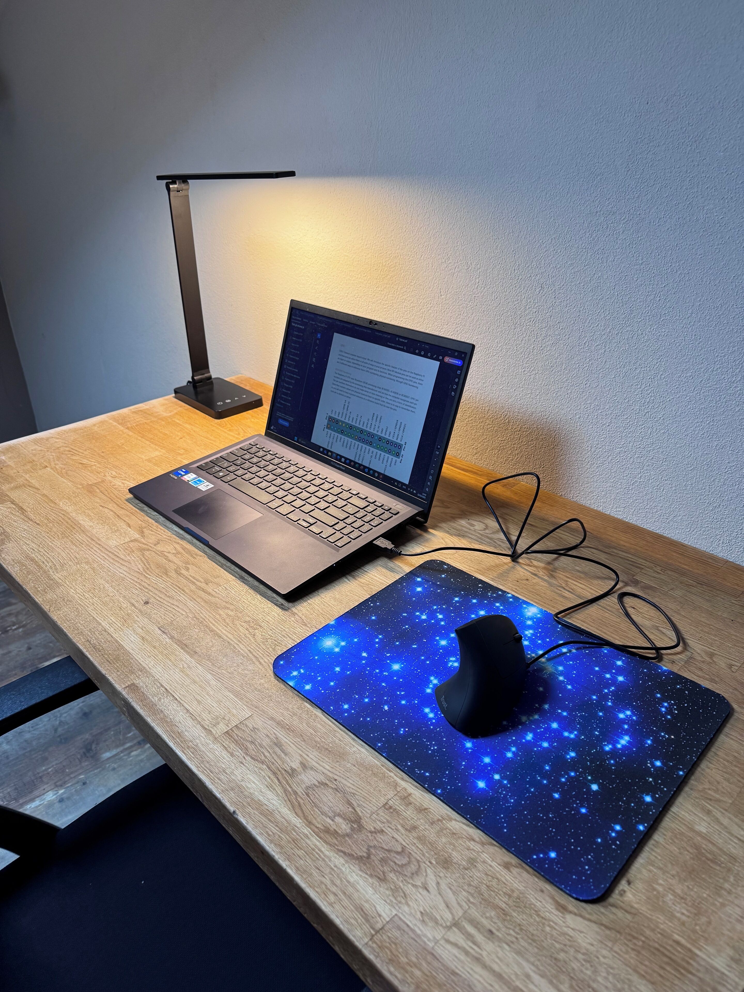 A laptop on a wooden desk with a starry mouse pad and a black lamp.