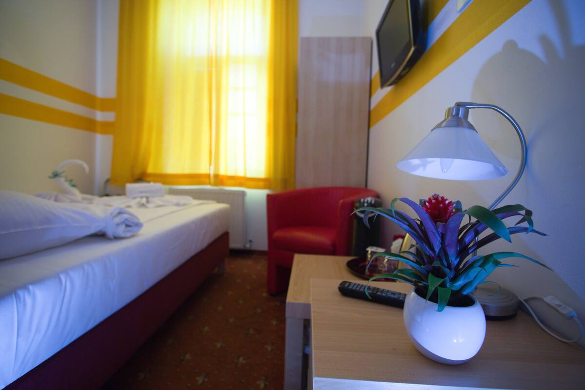 Une chambre d’hôtel avec un lit, une chaise rouge, un bureau avec une lampe, un vase de fleurs et une télécommande.