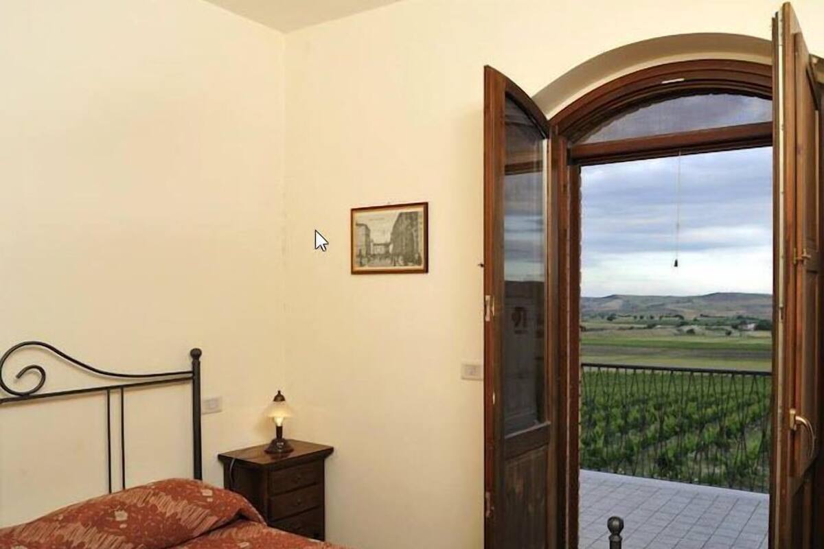 A bedroom with a bed, a nightstand with a lamp, a framed picture on the wall, and a balcony with a view of the countryside.