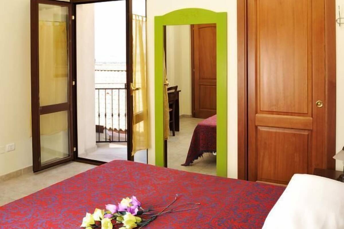 A hotel room with a bed, a green archway, a wooden door, and a balcony with a railing.