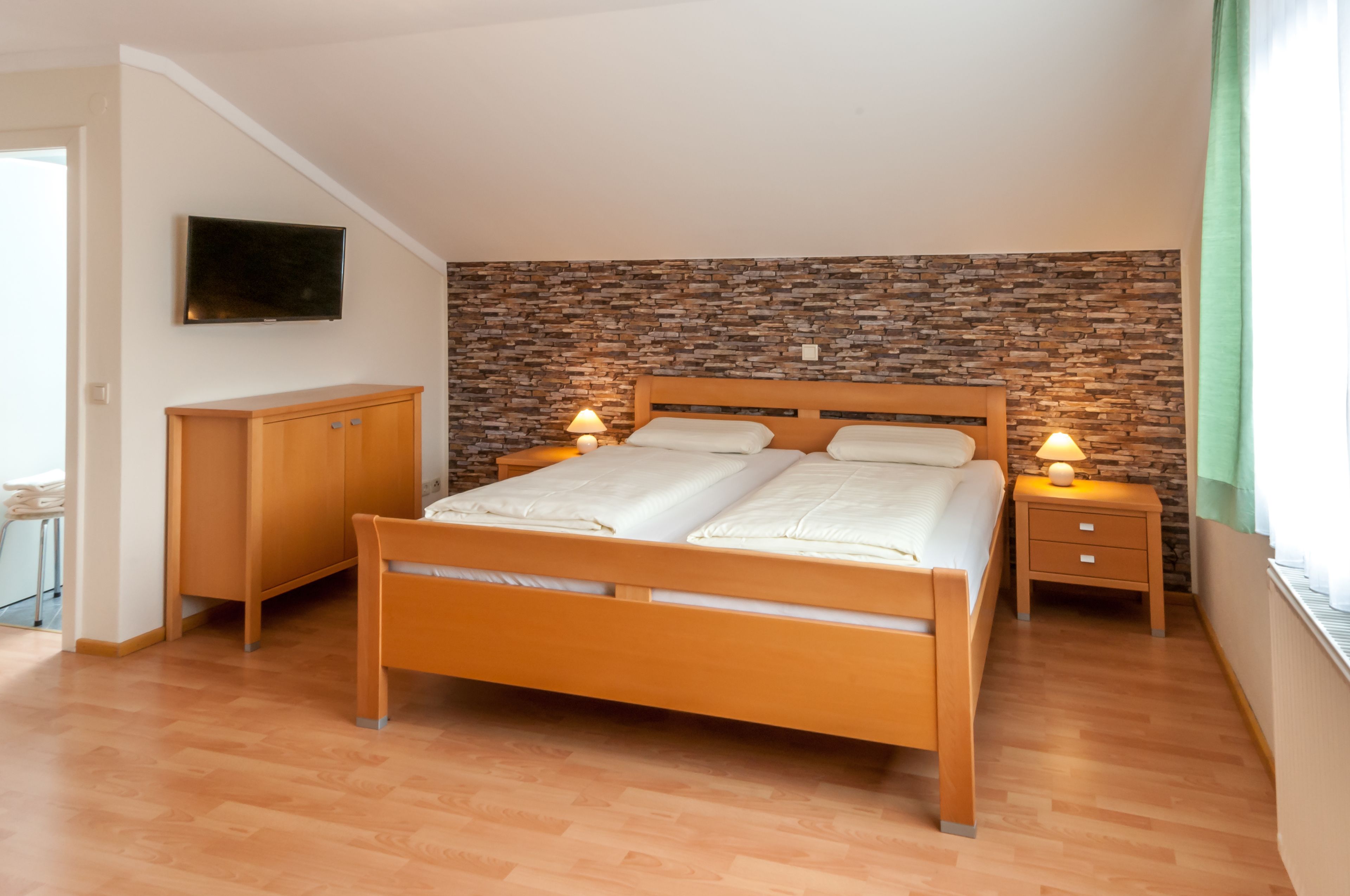 A bedroom with a wooden bed, two bedside tables with lamps, a flat-screen TV on the wall, and a stone-patterned accent wall.